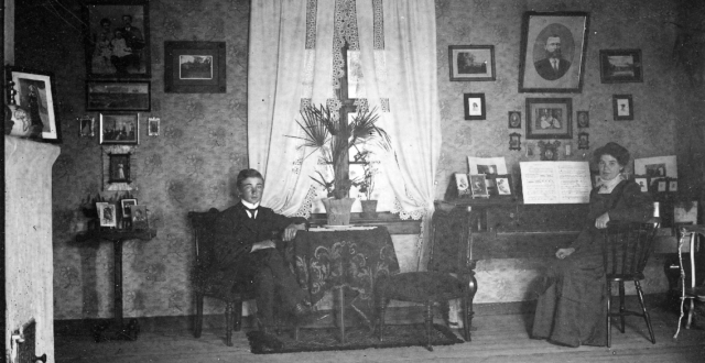 Puutarhuri Nilssonin pianosali Sippolassa v. 1912. Kuvassa nuori mies istuu pöydän ääressä , jossa on kukka pöydällä. Nainen istuu pianon ääressä. Seinällä on tauluja. Huoneessa on ikkuna, jossa valkoiset verhot.