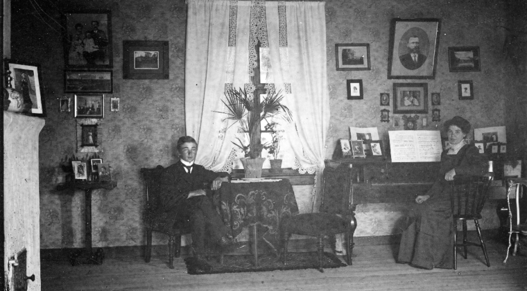 Puutarhuri Nilssonin pianosali Sippolassa v. 1912. Nuori mies istuu pöydän ääressä , jossa on kukka pöydällä. Nainen istuu pianon ääressä. Seinällä on tauluja. Huoneessa on ikkuna, jossa valkoiset verhot.