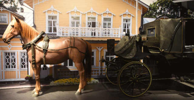 Mustilan kartanon kuomuvaunut, joita vetää ruskea hevonen. Taustalla keltainen kartanorakennus.