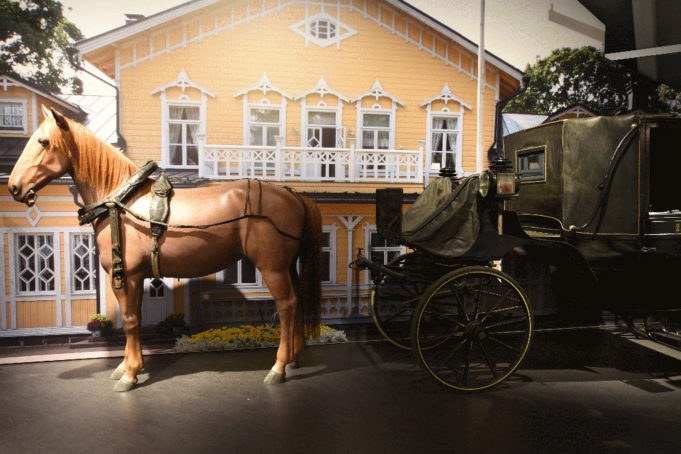 Mustilan kartanon kuomuvaunut, joita vetää ruskea hevonen. Taustalla keltainen kartanorakennus.