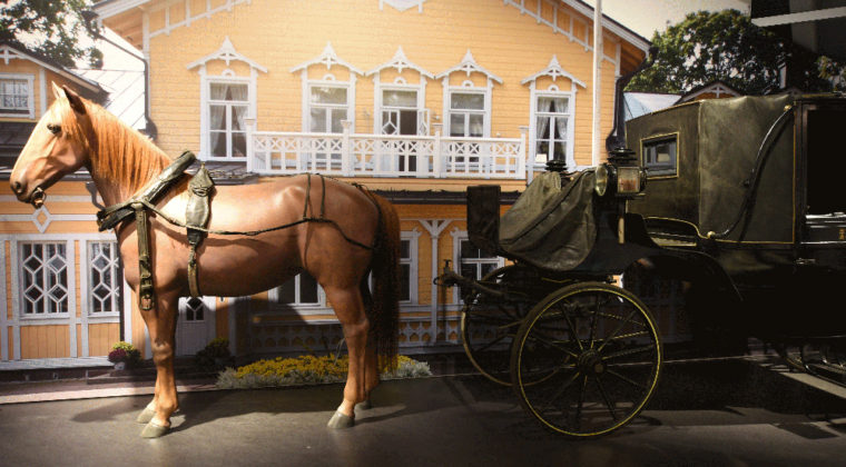 Mustilan kartanon kuomuvaunut, joita vetää ruskea hevonen. Taustalla keltainen kartanorakennus.