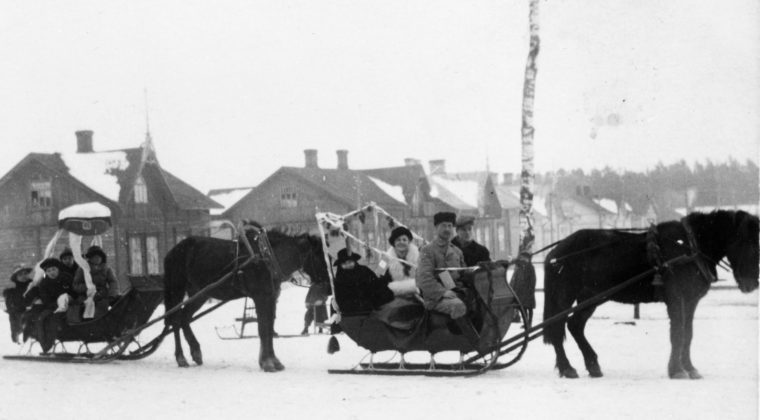 Penkinpainajaiset Kouvolassa talvella 1921. Kaksi rekeä, joita hevoset vetävät. Kyydissä abiturientteja. Toisessa reessa iso yliopppilashattu koristeena. Taustalla Kouvolan torilla olevia rakennuksia.
