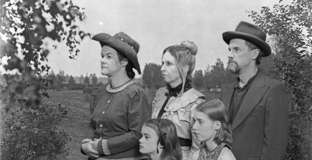 Ryhmäkuva, jossa kaksi naista ja mies sekä kaksi tyttöä puutarhamaiseman edessä seisomassa entisajan vaatteissa.