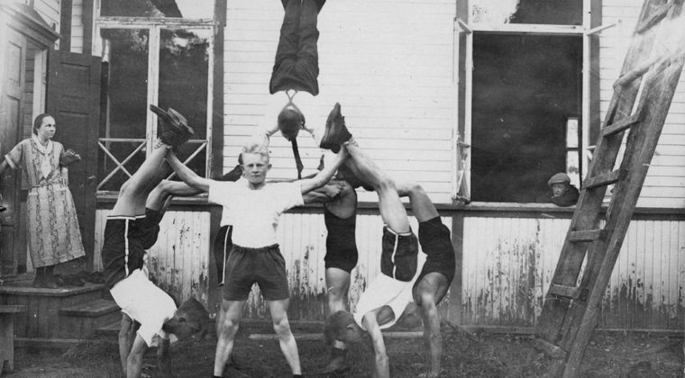 Kahdeksan Kuusankosken Puhdin poikaa harjoittelemassa pyramidin tekoa Kuusankosken Työväentalon edustalla. kesällä 1921. Nainen katsoo vieressä esitystä ja poika ikkunan takaa.
