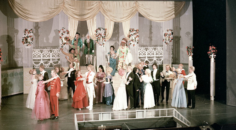 Lukuisia näyttelijöitä hienoissa puvuissa harjoittelemassa tanssikohtausta Kouvolan teatterin näyttämöllä.