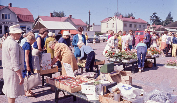 Kesäisessä värikuvassa torikauppiaita myymässä asiakkaille perunaa, mansikkaa, herneitä ja kukkia. Etualan myyntipöydässä perunalaatikko, hernelaatikko sekä kaksi mansikkalaatikkoa.