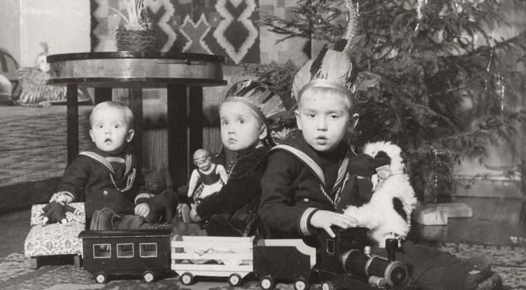 kolme lasta leikkimässä joulukuusen vieressä joululahjoilla. Lasten edessä pyörillä kulkeva vedettä juna ja sen vaunuja. Lapsilla käsissä nukkeja sekä pehmolelu.