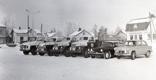 Kuvan etualalla rivissä kaksi Kouvoaln autokoulun henkilöautoa sekä neljä kuorma-autoa. Taustalla puurakennuksia.