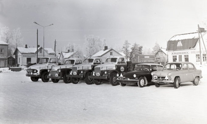 Kuvan etualalla rivissä kaksi Kouvoaln autokoulun henkilöautoa sekä neljä kuorma-autoa. Taustalla puurakennuksia.
