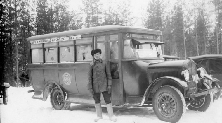 Linja-auto pysähtyneenä. Mies seisomassa linja-auton oviaukon edessä. Maassa on lunta.