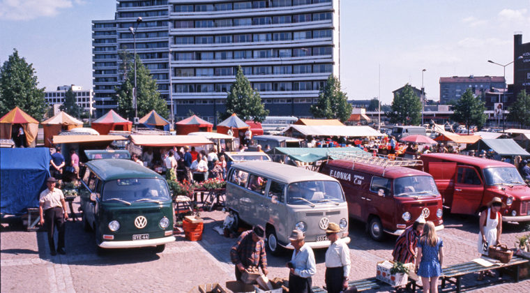 Värikuvassa etualalla torikauppiaita ja asiakkaita myyntipöydän ympärillä. Heidän takanaan vihreän, harmaan ja punaisen väriset Volkswagen pakettiautot sekä punainen Ford-pakettiauto. Takana torikojuja sekä Torikadun tornitalot ja kaupungintalo.