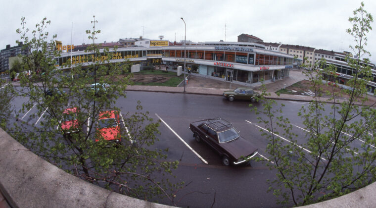 Etualalla autoja parkissa pysäköintiruuduissa. Takana kolme Hansakeskuksen neliönmuotoista liikerakennusta. Vasemmassa reunassa olevassa rakennuksessa toimi mm. Heps-tavararatalo.