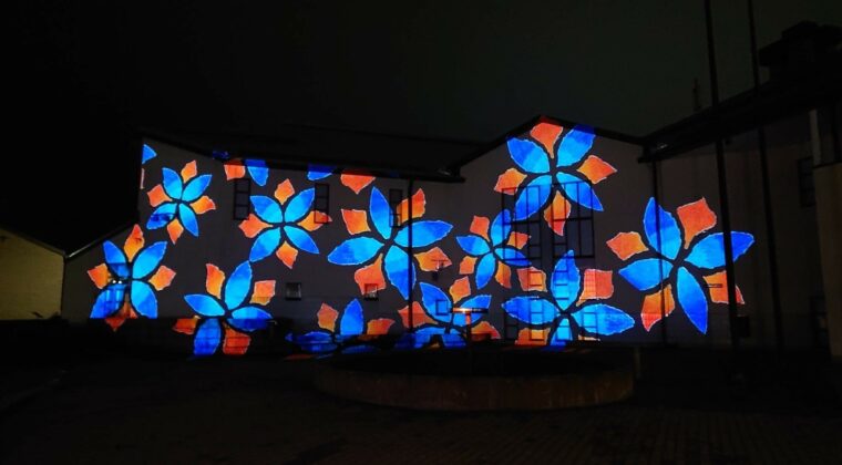 Valo heijastettuna Kouvolatalon sisäpihalla rakennuksen seinään. Sinisiä kukkakuvioita ja oranssit lehdet.