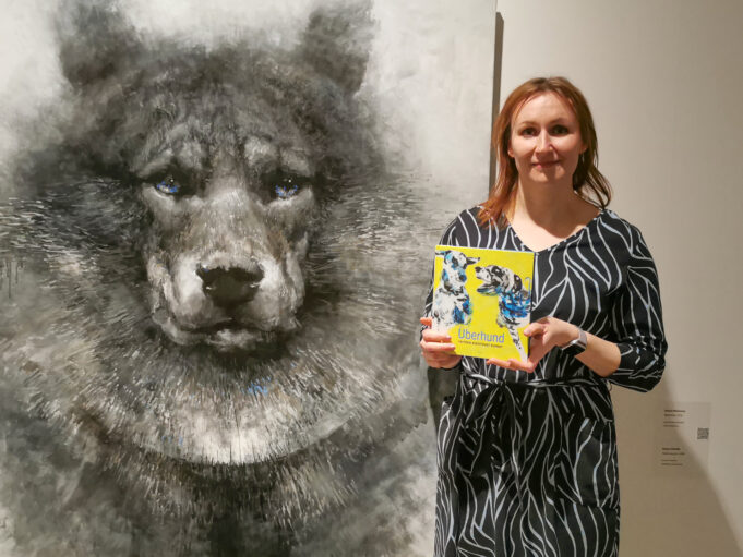 Kuvassa nainen koira-aiheisen taideteoksen vieressä. Kädessä keltakantinen kirja.