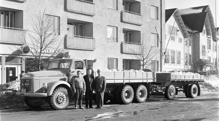 Kolme miestä seisoo kadulla Volvo-merkkisen kuorma-auton edessä. Takana Salpausselänkatu 34 kerrostalo, jonka katutasossa Autosalpa Oy:n liike. Puulyseon edustalla paljon polkupyöriä. Kuvattu keväällä, kadulla lammikoita ja kadun reunassa lumikinoksia.