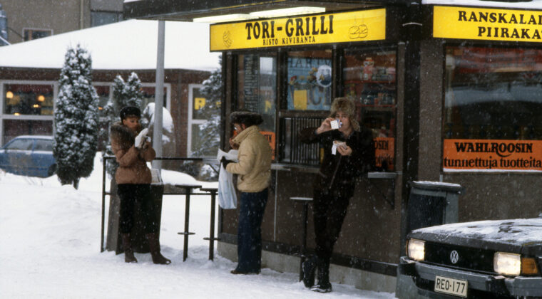 Kaksi miestä ja nainen tori-grillin edustalla talvella syömässä kioskista ostettuja tuotteita. Lunta sataa, miehillä on päässä karvahatut. Etualalla Volskwagen-henkilöauton keulaa.