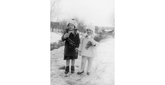 Kaksi pientä tyttöä vierekkäin talvivaatteissa loskaisella kevättiellä. Molemmilla tytöillä pajunkissakimput kainalossaan. Taustalla näkyy Voikkaan rakennuksia.