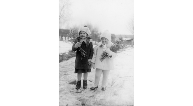 Kaksi pientä tyttöä vierekkäin talvivaatteissa loskaisella kevättiellä. Molemmilla tytöillä pajunkissakimput kainalossaan. Taustalla näkyy Voikkaan rakennuksia.