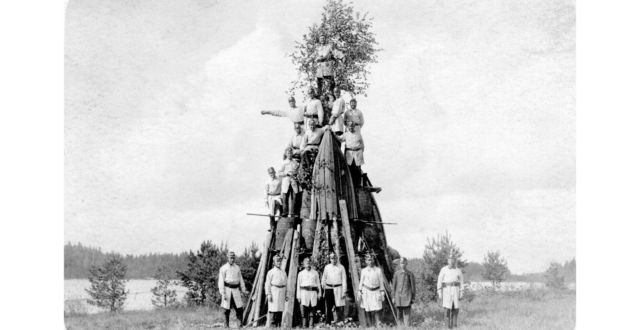 Voikkaan tehtaan palokunnan miehiä kokoontuneina juhannuskokon edustalle palopuvuissaan. Seitsemän miestä seisoo kokon edessä. Kokon päälle on kiivennyt kymmenen palomiestä eri korkeuksille, joista suurin osa seisoo tynnyrien päällä. Kokon latvassa on juhannuskoivu.