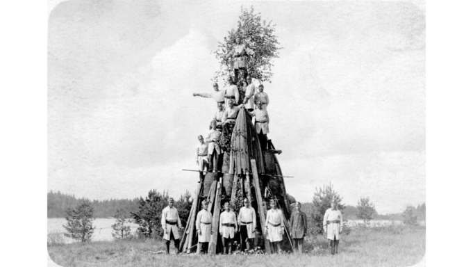 Voikkaan tehtaan palokunnan miehiä kokoontuneina juhannuskokon edustalle palopuvuissaan. Seitsemän miestä seisoo kokon edessä. Kokon päälle on kiivennyt kymmenen palomiestä eri korkeuksille, joista suurin osa seisoo tynnyrien päällä. Kokon latvassa on juhannuskoivu.