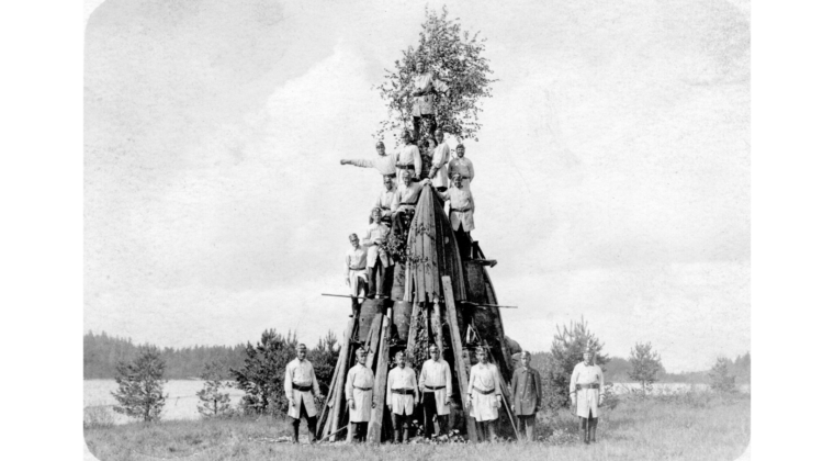 Voikkaan tehtaan palokunnan miehiä kokoontuneina juhannuskokon edustalle palopuvuissaan. Seitsemän miestä seisoo kokon edessä. Kokon päälle on kiivennyt kymmenen palomiestä eri korkeuksille, joista suurin osa seisoo tynnyrien päällä. Kokon latvassa on juhannuskoivu.