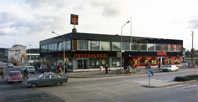 Näkymä Valtakadun yli kaksikerroksiseen liikerakennukseen. Autoja, pyöräilijöitä ja jalankulkijoita kulkee rakennuksen edustalla. Rakennuksen katolla Osuuspankin kyltti. Osuuspankin konttori sijaitsee rakennuksen katutasossa Valtakadun ja Keskusaukion kulmauksessa. Sen yläpuolella toisessa kerroksessa kampaamo ja parturi. Rakennuksen toisessa päässä Valtakadulla on tavaratalo Halpis sekä yläkerrassa PK-kahvila. Rakennuksen takana vasemmalla näkyy Shellin ja Esson huoltoasemien kyltit sekä Pienteollisuustalo.