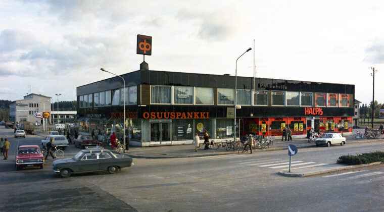 Näkymä Valtakadun yli kaksikerroksiseen liikerakennukseen. Autoja, pyöräilijöitä ja jalankulkijoita kulkee rakennuksen edustalla. Rakennuksen katolla Osuuspankin kyltti. Osuuspankin konttori sijaitsee rakennuksen katutasossa Valtakadun ja Keskusaukion kulmauksessa. Sen yläpuolella toisessa kerroksessa kampaamo ja parturi. Rakennuksen toisessa päässä Valtakadulla on tavaratalo Halpis sekä yläkerrassa PK-kahvila. Rakennuksen takana vasemmalla näkyy Shellin ja Esson huoltoasemien kyltit sekä Pienteollisuustalo.