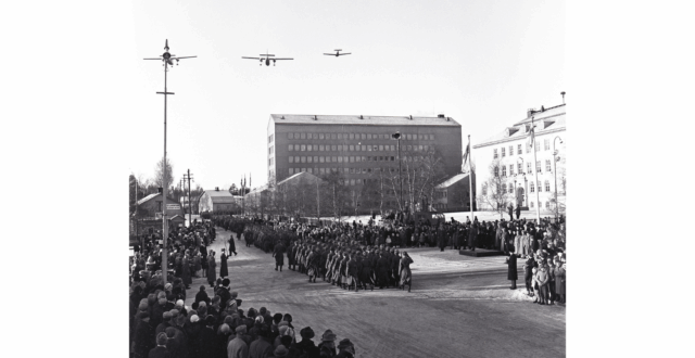 Sotilaita marssimassa paraatin vastaanottolavan edessä Kymen Lukon edustalla Salpausselänkadulla. Takana näkyy Kankaan koulu sekä Kymen lääninvirastotalo. Yleisöä seuraamassa kulkuetta kadun molemmilla puolilla. Kaksi ilmavoimien lentokonetta lentää kulkueen yläpuolella.
