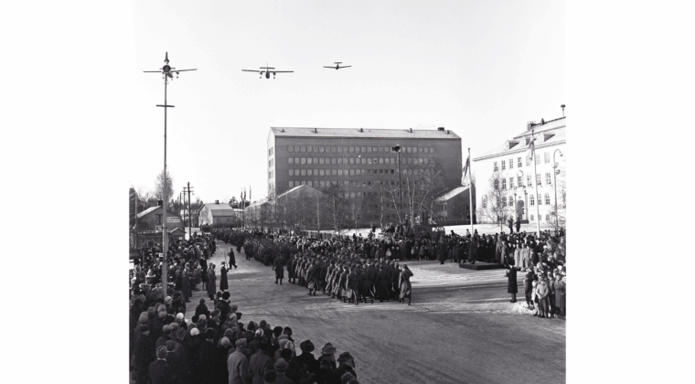 Sotilaita marssimassa paraatin vastaanottolavan edessä Kymen Lukon edustalla Salpausselänkadulla. Takana näkyy Kankaan koulu sekä Kymen lääninvirastotalo. Yleisöä seuraamassa kulkuetta kadun molemmilla puolilla. Kaksi ilmavoimien lentokonetta lentää kulkueen yläpuolella.