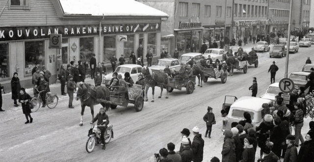 Talvisessa kuvassa abiturientteja istumassa hevosten vetämissä vaunuissa puisen Kouvolan Rakennusaineen talon edustalla. Yleisöä katsomassa kulkuetta kadun molemmilla puolilla. Henkilöautoja pysäköitynä kadun varteen. Kulkueen edessä etualalla moottoripyöräilijä. Takana Laukkarisen liiketalon vieressä näkyy Kauppalankatu 10-12 liikerakennukset.