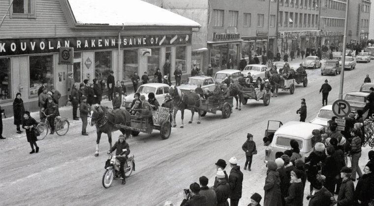 Talvisessa kuvassa abiturientteja istumassa hevosten vetämissä vaunuissa puisen Kouvolan Rakennusaineen talon edustalla. Yleisöä katsomassa kulkuetta kadun molemmilla puolilla. Henkilöautoja pysäköitynä kadun varteen. Kulkueen edessä etualalla moottoripyöräilijä. Takana Laukkarisen liiketalon vieressä näkyy Kauppalankatu 10-12 liikerakennukset.