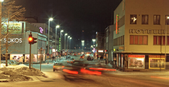 Autoja kulkemassa Kauppalankatua iltavalaistuksessa rautatieaseman suuntaan. Etualalla vasemmalla liikennevalot, jotka keltaisella. Liikennevalojen takana näkyy Sokoksen tavaratalon katutason näyteikkunoita. Sokosken takana näkyy Säästöpankin rakennus sekä Valioasun liikkeen kyltti. Oikealla keltainen Hotelli Kouvolan rakennus, jonka katutasossa näkyy Kausi-Kukka -liikkeen näyteikkunat. Takana näkyy Kouvolan rautatieasema. Maassa on lunta.