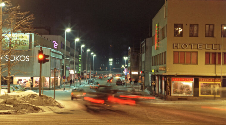 Autoja kulkemassa Kauppalankatua iltavalaistuksessa rautatieaseman suuntaan. Etualalla vasemmalla liikennevalot, jotka keltaisella. Liikennevalojen takana näkyy Sokoksen tavaratalon katutason näyteikkunoita. Sokosken takana näkyy Säästöpankin rakennus sekä Valioasun liikkeen kyltti. Oikealla keltainen Hotelli Kouvolan rakennus, jonka katutasossa näkyy Kausi-Kukka -liikkeen näyteikkunat. Takana näkyy Kouvolan rautatieasema. Maassa on lunta.