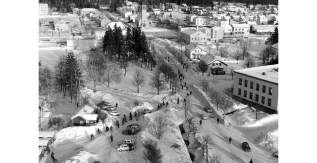 Etualalla autoja tehtaan pysäköintialueella. Polkupyöräilijöitä ja jalankulkijoita menossa tehtaalta keskustan suuntaan. Seuratalon takana Myllykosken tori ja kirkko sekä Myllykoskentien ja Kenraalintien liikerakennuksia. Maassa on paljon lunta.