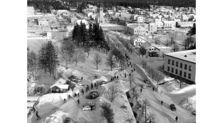 Etualalla autoja tehtaan pysäköintialueella. Polkupyöräilijöitä ja jalankulkijoita menossa tehtaalta keskustan suuntaan. Seuratalon takana Myllykosken tori ja kirkko sekä Myllykoskentien ja Kenraalintien liikerakennuksia. Maassa on paljon lunta.