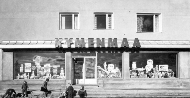 Mustavalkoisessa kuvassa etualalla viisi lasta kerrostalossa sijaitsevan myymälän edustalla. Kolme lasta leikkii pyörätelineissä, yksi lapsista seisoo lastenrattaiden vieressä ja yksi lapsi istuu myymälän portailla. Lasten takana näkyy katutasossa olevan myymälän näyteikkunoita, joissa näkyy mainossomisteita ja tuotteita mm. OTK:n kahvimainoksia. Ylhäällä näkyy kerrostalon asuntojen ikkunoita.