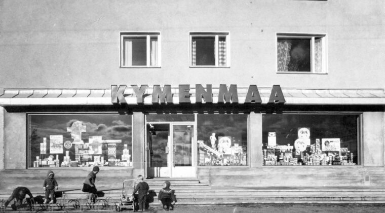 Mustavalkoisessa kuvassa etualalla viisi lasta kerrostalossa sijaitsevan myymälän edustalla. Kolme lasta leikkii pyörätelineissä, yksi lapsista seisoo lastenrattaiden vieressä ja yksi lapsi istuu myymälän portailla. Lasten takana näkyy katutasossa olevan myymälän näyteikkunoita, joissa näkyy mainossomisteita ja tuotteita mm. OTK:n kahvimainoksia. Ylhäällä näkyy kerrostalon asuntojen ikkunoita.