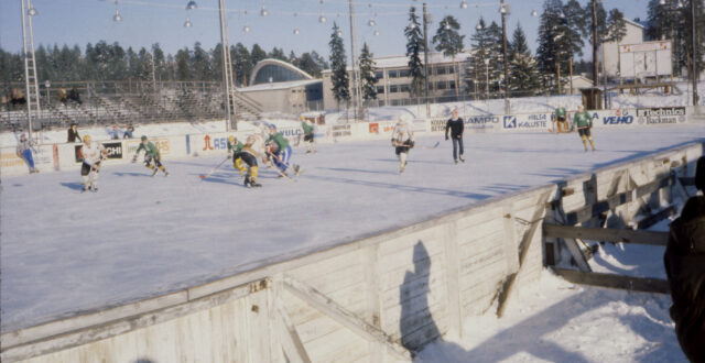 Kaukalon laidalta otettu kuva jääkiekkoilijoista, joista KooKoon pelaajilla musta-oranssi-valkoinen pelisasu. Vastustajan peliasussa pääväri vihreä ja musta. Tuomarilla musta vaatetus ja pipo päässä. Kaukalon toisella puolella näkyy puukatsomo. Takana näkyy Urheilupuiston koulu sekä Kouvolan uimahalli.