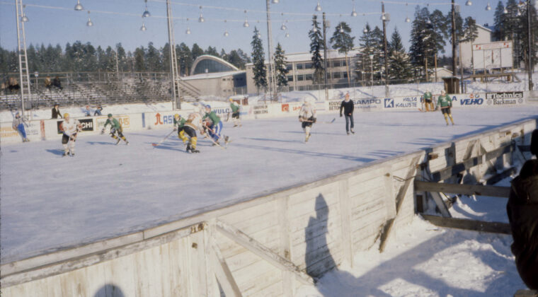 Kaukalon laidalta otettu kuva jääkiekkoilijoista, joista KooKoon pelaajilla musta-oranssi-valkoinen pelisasu. Vastustajan peliasussa pääväri vihreä ja musta. Tuomarilla musta vaatetus ja pipo päässä. Kaukalon toisella puolella näkyy puukatsomo. Takana näkyy Urheilupuiston koulu sekä Kouvolan uimahalli.
