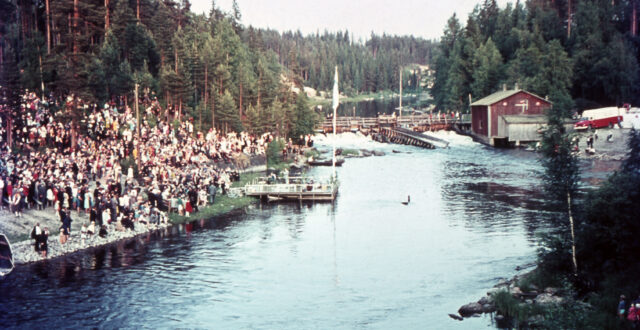 Värikuvassa runsaasti ihmisiä kokoontuneena joen rantaan, jossa lavalla menossa musiikkiesitys. Lavalla lipputangossa Suomen lippu. Oikealla näkyy Jokelan vanha mylly, jonka vieressä ihmisiä kulkemassa sillalla.