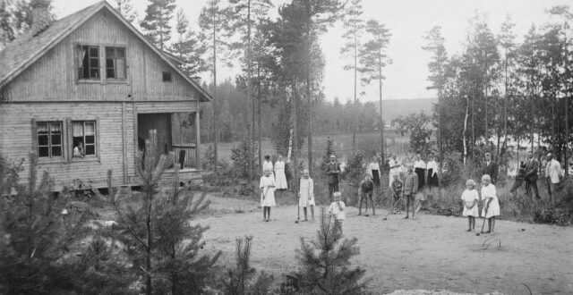 Etualalla hiekkakenttä, jossa neljä tyttöä ja neljä poikaa pelaamassa krokettia. Osalla on käsissään krokettimaila. Lasten takana nurmikolla kymmenen aikuista sekä koira. Käyrälampi näkyy takana. Hirsirakenteinen Ketomaan huvila näkyy vasemmalla, jonka ikkunasta katsoo nainen.