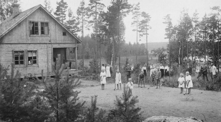 Etualalla hiekkakenttä, jossa neljä tyttöä ja neljä poikaa pelaamassa krokettia. Osalla on käsissään krokettimaila. Lasten takana nurmikolla kymmenen aikuista sekä koira. Käyrälampi näkyy takana. Hirsirakenteinen Ketomaan huvila näkyy vasemmalla, jonka ikkunasta katsoo nainen.