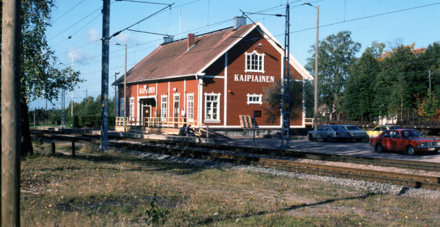 Värikuvassa etualalla kulkee kaksi junaraidetta. Raiteiden takana asemarakennus, jossa päädyssä ja sivulla valkoisella teksti: KAIPIAINEN. Kuva on otettu kaakon suunnalta. Kaksi miestä kunnostamassa asemarakennuksen edustalla olevaa puista kaidetta. Kolmas mies nojaa kaiteeseen. Rakennuksen edustalla oikealla neljä henkilöautoa pysäköitynä. Sää on aurinkoinen, puissa näkyy hieman ruskan merkkejä.
