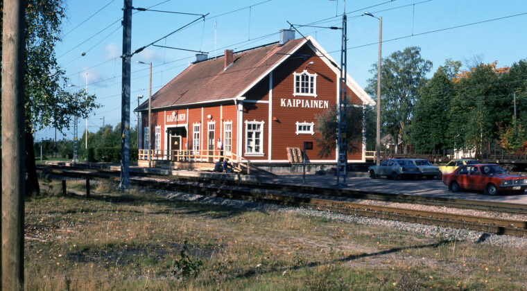 Värikuvassa etualalla kulkee kaksi junaraidetta. Raiteiden takana asemarakennus, jossa päädyssä ja sivulla valkoisella teksti: KAIPIAINEN. Kuva on otettu kaakon suunnalta. Kaksi miestä kunnostamassa asemarakennuksen edustalla olevaa puista kaidetta. Kolmas mies nojaa kaiteeseen. Rakennuksen edustalla oikealla neljä henkilöautoa pysäköitynä. Sää on aurinkoinen, puissa näkyy hieman ruskan merkkejä.