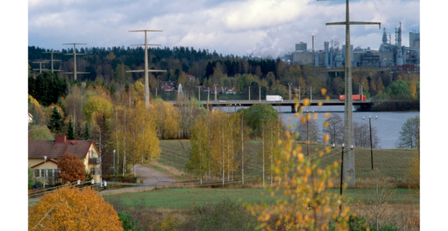 Näkymä betonipylväistä Mäkikylän suunnasta kuvattuna. Etualalla oikealla yksi betonipylväs, jonka takana näkyy Kymijokea, Kuusaan silta sekä Kuusanniemen tehdas. Vasemmalla näkyy neljä pylvästä. Etualalla vasemmalla keltainen omakotitalo. Puissa on ruskan värejä.