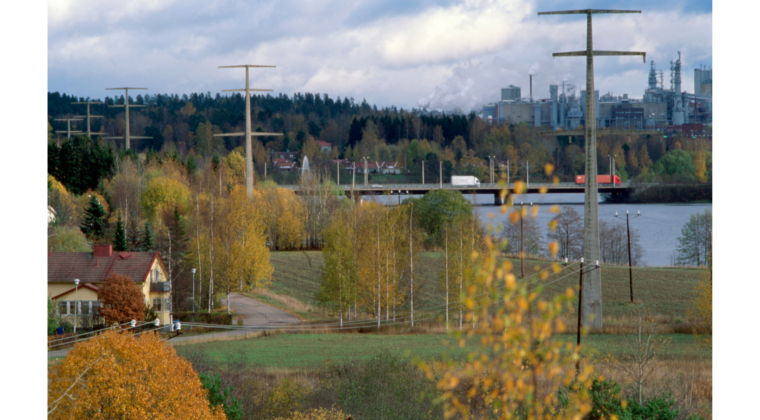 Näkymä betonipylväistä Mäkikylän suunnasta kuvattuna. Etualalla oikealla yksi betonipylväs, jonka takana näkyy Kymijokea, Kuusaan silta sekä Kuusanniemen tehdas. Vasemmalla näkyy neljä pylvästä. Etualalla vasemmalla keltainen omakotitalo. Puissa on ruskan värejä.